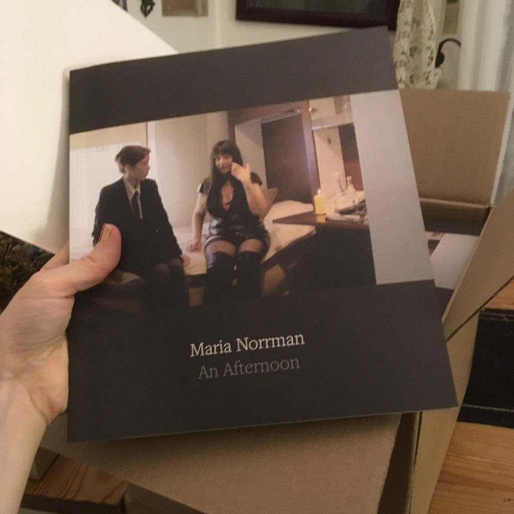 A hand holding the artist book An Afternoon by Maria Norrman. 
The cover displays two persons on a bed, one younger wearing a suit and tie, one older in black lingerie. 