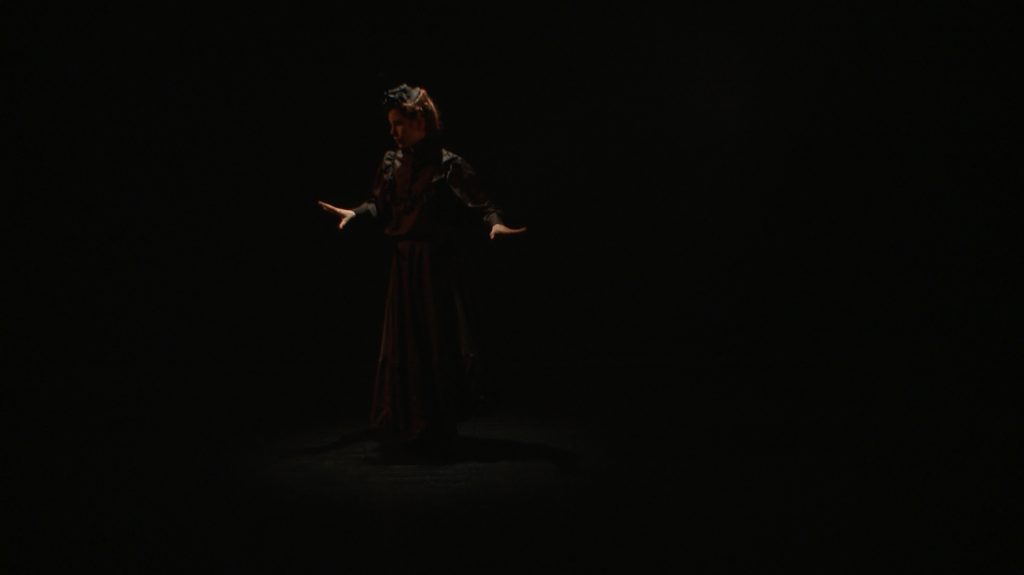 Video still from artwork where artist Maria Norrman interprets and invokes dance artist Jane Avril.
The artist in a dancing pose, dressed in black 19th century clothing.