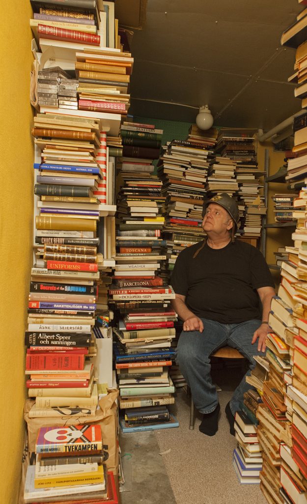 The photo work Burden of Knowledge. 
A seated man with a helmet sits in a room with crammed bookshelves and book piles, from top to bottom.